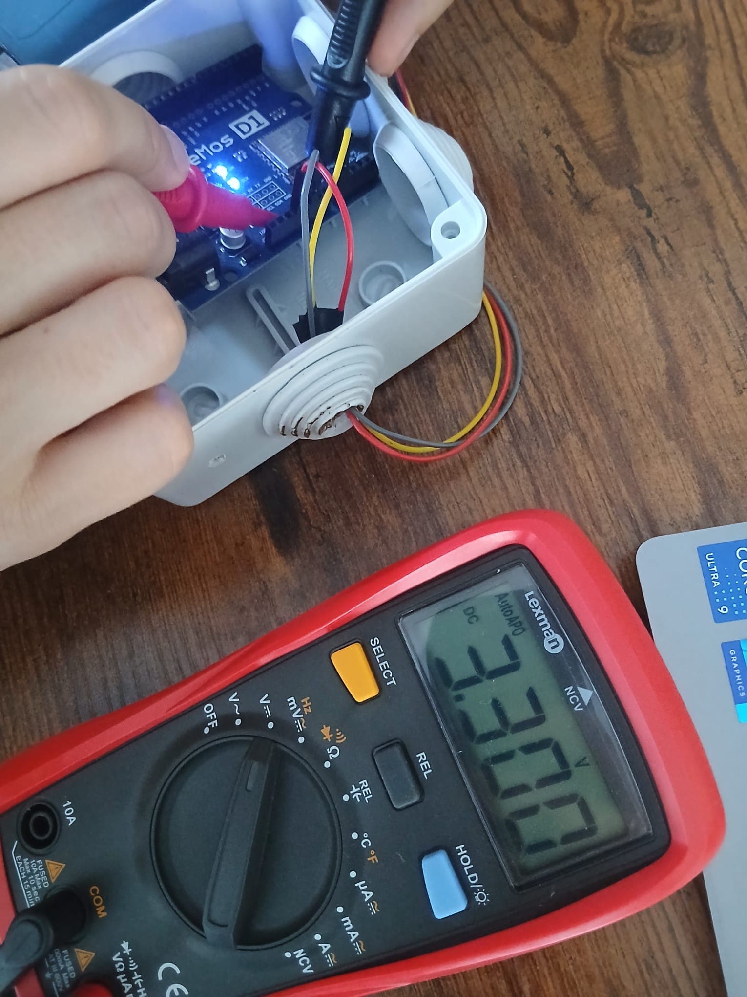 Multimeter probes on the 3V3 and GND pins of the Wemos inside its weatherproof box, display showing 3.30 V.