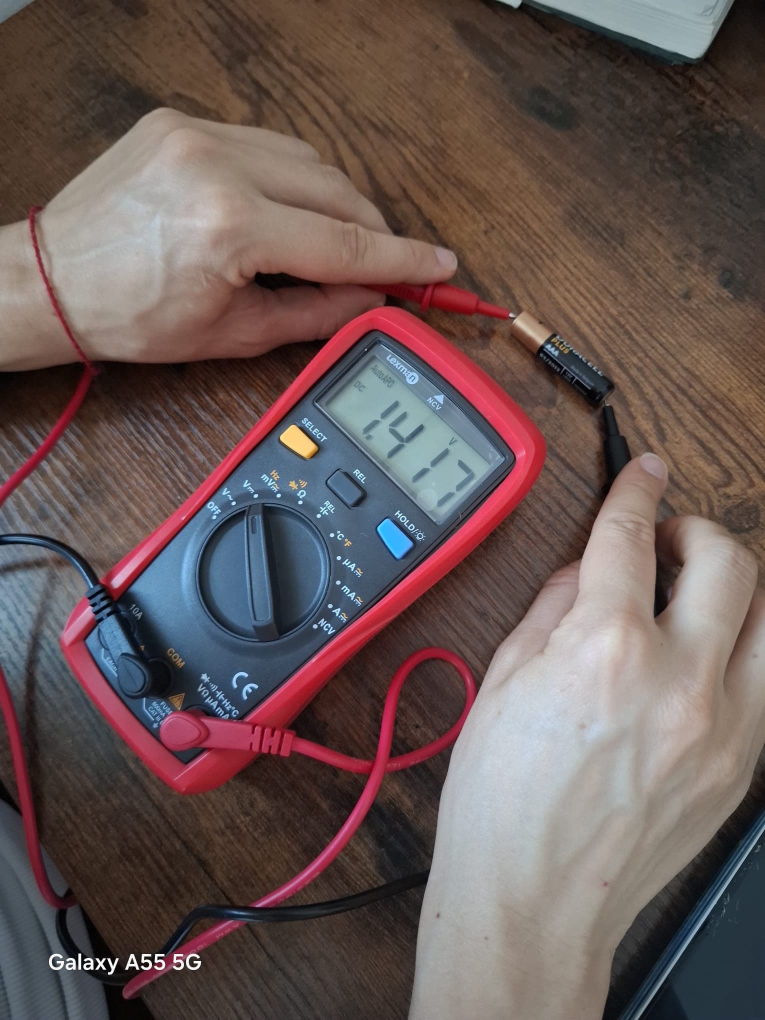 Multimeter with the red probe on the positive end and the black probe on the negative end of a AAA battery, display reading about 1.4 V.
