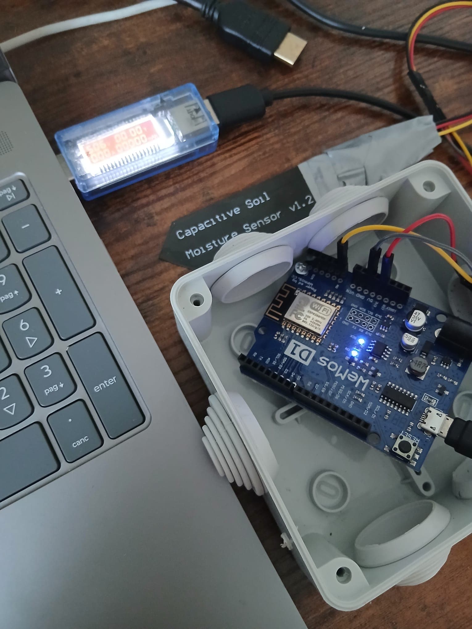 The Wemos in its weatherproof box connected to the laptop through the USB power meter, with the capacitive soil-moisture sensor lying next to it.