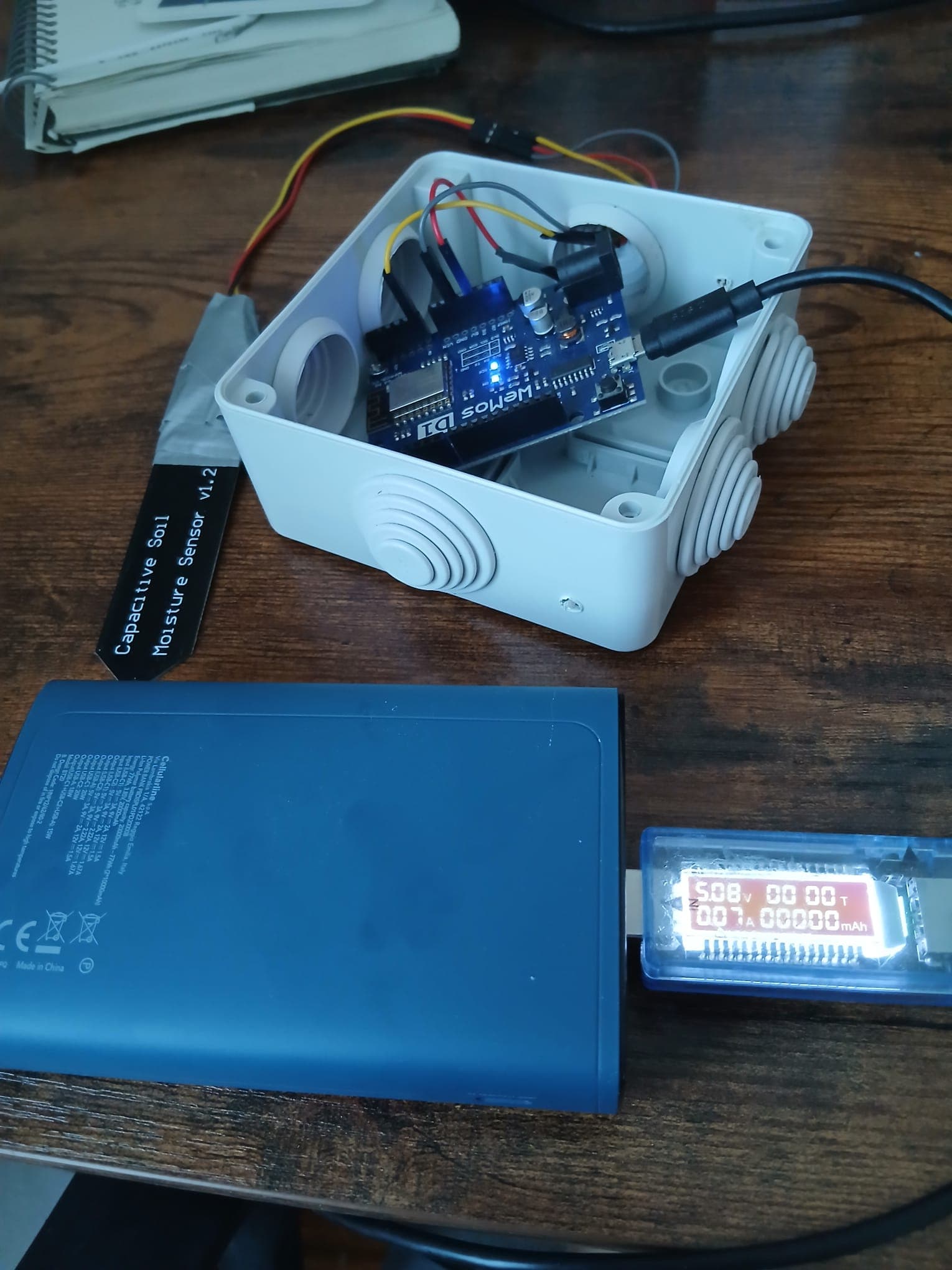 Power bank connected to the USB power meter and to the Wemos in its weatherproof box, with the meter display lit and showing voltage and current.