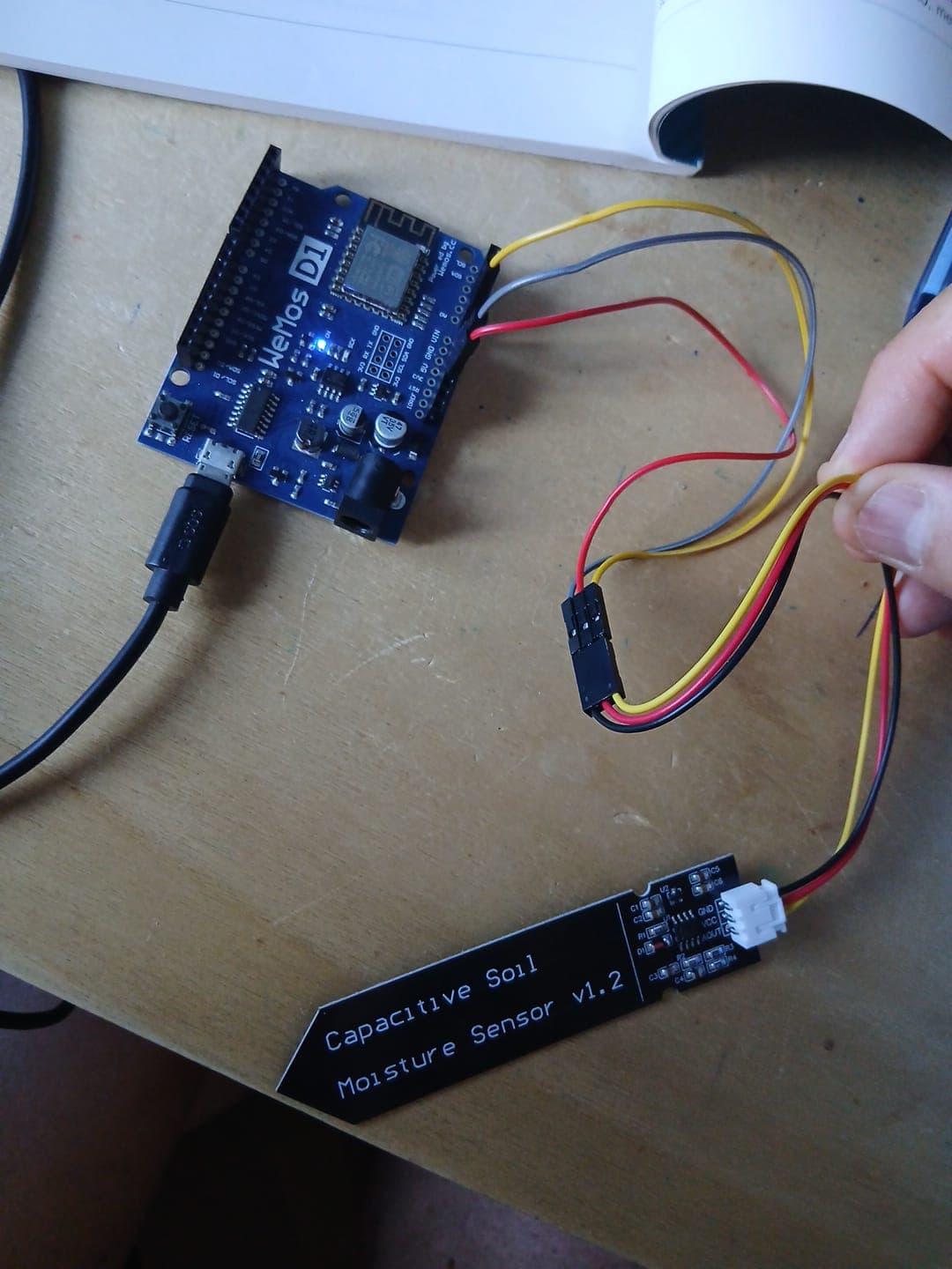 A Wemos board on the left connected by three jumper wires to a soil moisture sensor on the right, on a wooden desk.